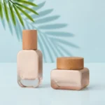 Glass lotion bottle and cream jar with natural wood closures on a light cyan tabletop with visible green palm foliage