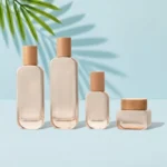 Three glass skincare bottles and a matching cream jar with cylindrical natural wood closures on a light cyan tabletop beneath visible green palm leaves