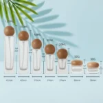 Dimension guide for clear glass lotion bottles and cream jars with rounded natural wood closures on a light cyan background with visible green palm leaves