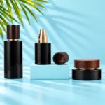 Two black glass bottles, exposed gold spray pump, detached wood cap and matching cream jar on cyan tabletop and rectangular platform with green palm leaves