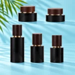 Three black glass bottles and two cream jars with dark wood closures arranged on matching light cyan platforms with visible green palm leaves