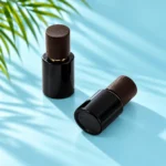 Two compact black glass bottles with dark wood caps, one upright and one reclining, on cyan tabletop beneath green palm leaves
