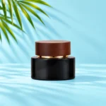Compact black glass cream jar with dark wood lid and gold collar on cyan tabletop beneath green palm leaves
