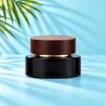 Glossy black glass cream jar with a flat dark wood lid and gold-tone collar on a light cyan tabletop beside green palm leaves