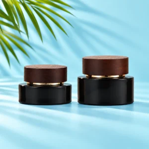 Two black glass cream jars with dark wood lids and gold collars on a cyan tabletop beneath visible green palm leaves