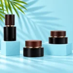 Two black glass cream jars and a matching bottle with dark wood closures on light cyan rectangular platforms beside green palm leaves