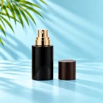 Compact black glass spray bottle with exposed gold atomizer and detached dark wood cap on cyan tabletop beneath green palm leaves