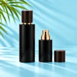 Tall capped black glass bottle and compact black spray bottle with gold atomizer and detached wood cap on a cyan tabletop beneath green palm foliage