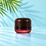 Close view of burgundy gradient glass cream jar and dark rounded wood lid on a light cyan tabletop with visible green palm leaves