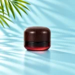 Compact burgundy gradient glass cream jar with a dark rounded wood lid on a light cyan tabletop with visible green palm leaves