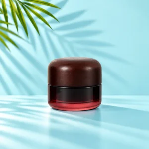 Medium profile burgundy gradient glass cream jar with a dark rounded wood lid on a light cyan tabletop with visible green palm leaves