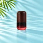 Compact burgundy gradient glass lotion bottle with a dark rounded wood cap and black pump option on a light cyan tabletop with visible palm foliage