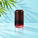 Compact burgundy gradient glass lotion bottle with a dark rounded wood cap and white pump option on a light cyan tabletop with visible palm foliage