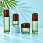 Three frosted green bottles and a matching cream jar with rounded dark wood closures on a light cyan cylinder and tabletop beside green palm leaves