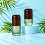 Two compact frosted green glass bottles with dark wood caps on a matching light cyan cylinder and tabletop with visible green palm leaves