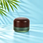 Compact frosted green glass cream jar with a rounded dark wood lid on a light cyan tabletop beside green palm foliage