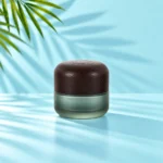 Medium-profile frosted green glass cream jar with a rounded dark wood lid on a light cyan tabletop beneath visible green palm leaves