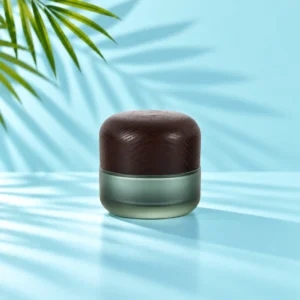Medium-profile frosted green glass cream jar with a rounded dark wood lid on a light cyan tabletop beneath visible green palm leaves
