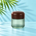 Tall-profile frosted green glass cream jar with a rounded dark wood lid on a light cyan tabletop beneath visible green palm leaves