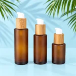 Three amber frosted glass lotion bottles with bamboo collars and white pumps on a pale blue cyan tabletop with green palm leaves