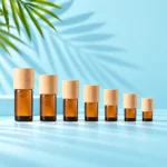 Seven compact amber glass essential oil bottles with light natural wood caps on a mint tabletop with a matching cyan backdrop and visible green palm leaves