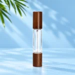 Clear double ended glass perfume bottle with dark wood caps on both ends on a pale blue cyan tabletop