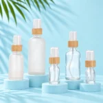 Five clear and frosted rounded shoulder glass bottles with bamboo collars and clear protective caps on matching pale blue cyan platforms
