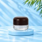 Compact clear glass cream jar with rounded dark solid wood lid on a cyan platform, nominal 30g version.