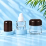 Open clear glass lotion bottle with white pump, detached wood cap resting on a cyan platform and matching cream jar.