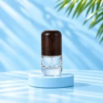 Compact clear glass lotion bottle with rounded dark wood cap on a cyan platform beneath green palm leaves.