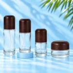 Three clear glass lotion bottles and a matching cream jar with rounded dark wood closures on a cyan tabletop.