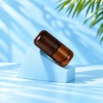 Compact amber glass lotion bottle tilted on a cyan support with rounded dark wood cap.