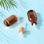 Frosted brown glass bottles with bamboo and sapele collar assemblies and a detached glass pipette resting on a pale blue tabletop.