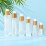 Six frosted clear glass lotion bottles with bamboo collars and white pumps on a pale blue cyan tabletop with green palm leaves