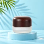 Compact frosted glass cream jar with a rounded dark wood lid on a cyan platform matching the mint tabletop and palm background