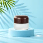 Medium frosted glass cream jar with a rounded dark wood lid on a cyan platform matching the mint tabletop and palm background