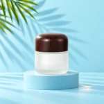 Tall frosted glass cream jar with a rounded dark wood lid on a cyan display platform matching the mint tabletop and green palm background