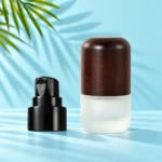 Compact frosted glass lotion bottle with a rounded dark wood cap beside a detached black pump on a mint tabletop