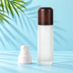 Tall frosted glass lotion bottle with a rounded dark wood cap beside a detached white pump in a mint palm studio