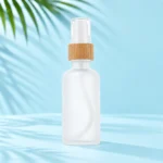 Frosted glass rounded shoulder bottle with a bamboo collar, white dispenser and clear protective cap on a pale blue cyan tabletop