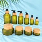 Six olive green frosted glass bottles and four matching bamboo lid cream jars in a size lineup on a pale blue cyan tabletop
