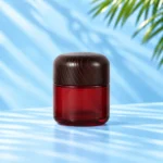 Red glass cream jar in the 100g nominal size with a rounded dark wood lid on a cyan tabletop.