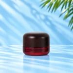 Compact red glass cream jar in the 30g nominal size with a rounded dark wood lid on a cyan tabletop.