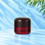 Red glass cream jar in the 50g nominal size with a rounded dark wood lid on a cyan tabletop.
