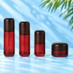 Three red glass lotion bottles and one matching red glass cream jar with rounded dark wood closures on a cyan tabletop.