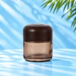 Smoke gray translucent glass cream jar in the 100g nominal size with a rounded dark wood lid on a cyan tabletop.