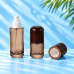 Two smoke gray glass lotion bottles, one with exposed white pump, with a detached wood cap resting on the cyan tabletop and showing its white insert.