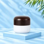 Opaque white cream jar with rounded dark solid wood lid on a pale blue cyan platform. Nominal 50g version.