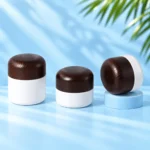 Three opaque white cream jars with rounded dark wood lids, shown upright and tilted on a cyan tabletop and pedestal.